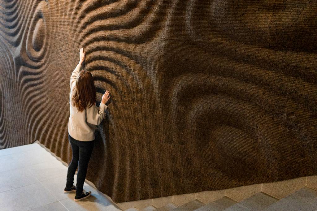 Mulher de costas passa a mão numa parede enrugada com a textura da cortiça