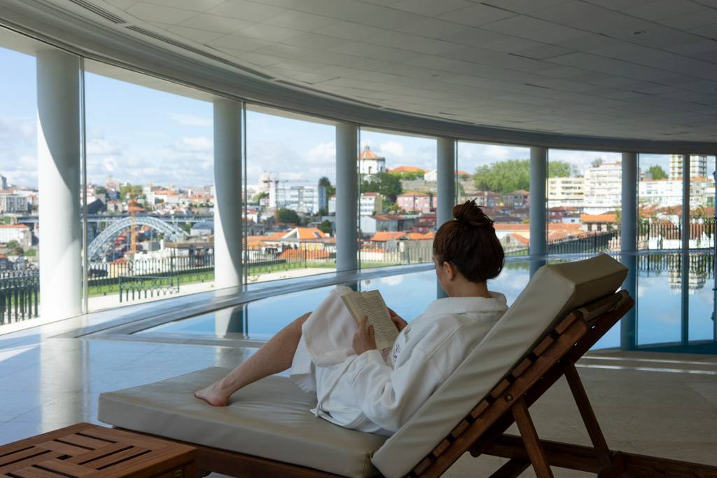 Mulher lendo um livro numa espreguiçadeira em frente à piscina interna, com vista da cidade do Porto