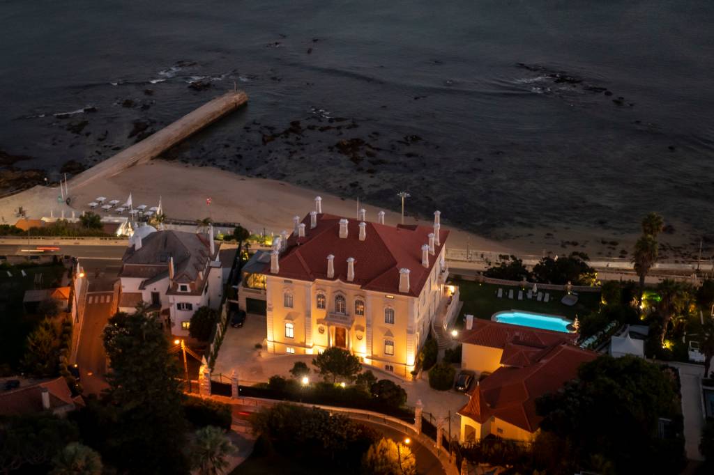 Vista aérea de um palacete amarelo com piscina à beira-mar, ao anoitecer