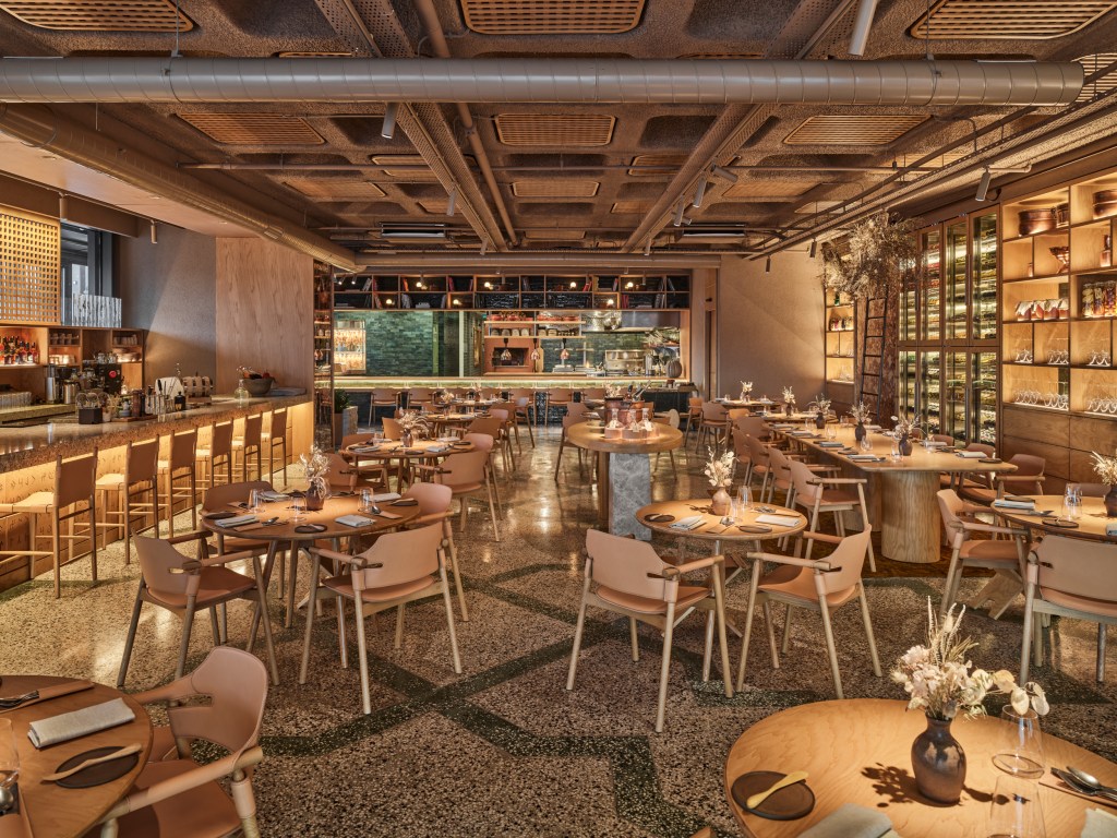 A imagem colorida mostra o salão de um restaurante com muita madeira e cimento.