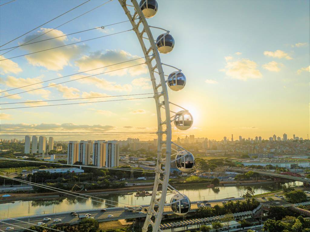 A fotografia colorida mostra as cabines de uma roda-gigante no fim do dia, com o céu azul e amarelo