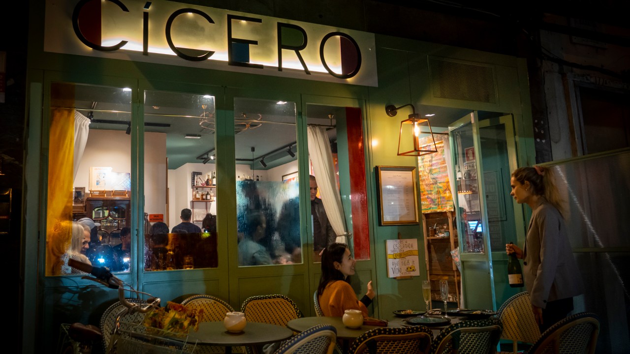 Fachada do restaurante Cícero com detalhes em madeira azul, mesas redondas e cadeiras e, no fundo, um letreiro luminoso com o nome do restaurante