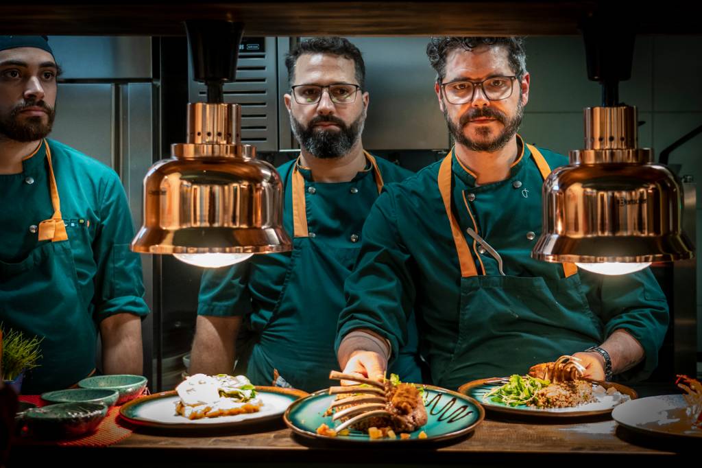 Janela da cozinha do restaurante onde se vê três cozinheiros vestidos de verde, mostrando os pratos