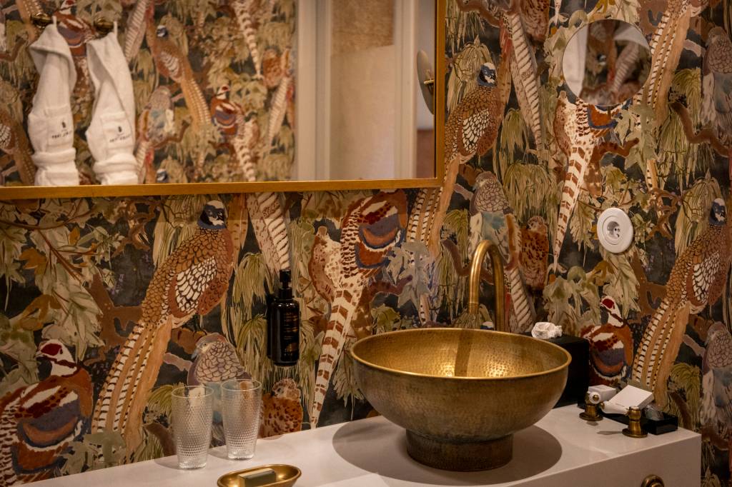 detalhe da pia do banheiro do quarto do hotel: cuba e torneiras douradas, espelho e um papel de parede com pássaros exóticos