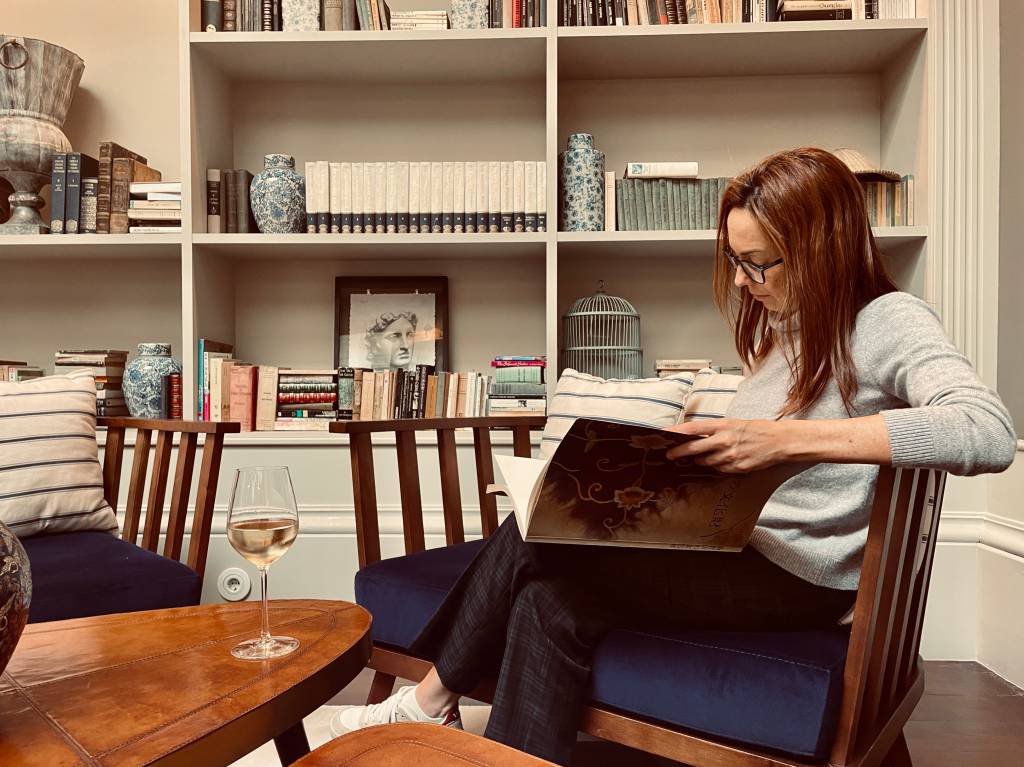 Mulher folheando um livro sentada numa poltrona, com um copo de vinho branco apoiado na mesinha em frente