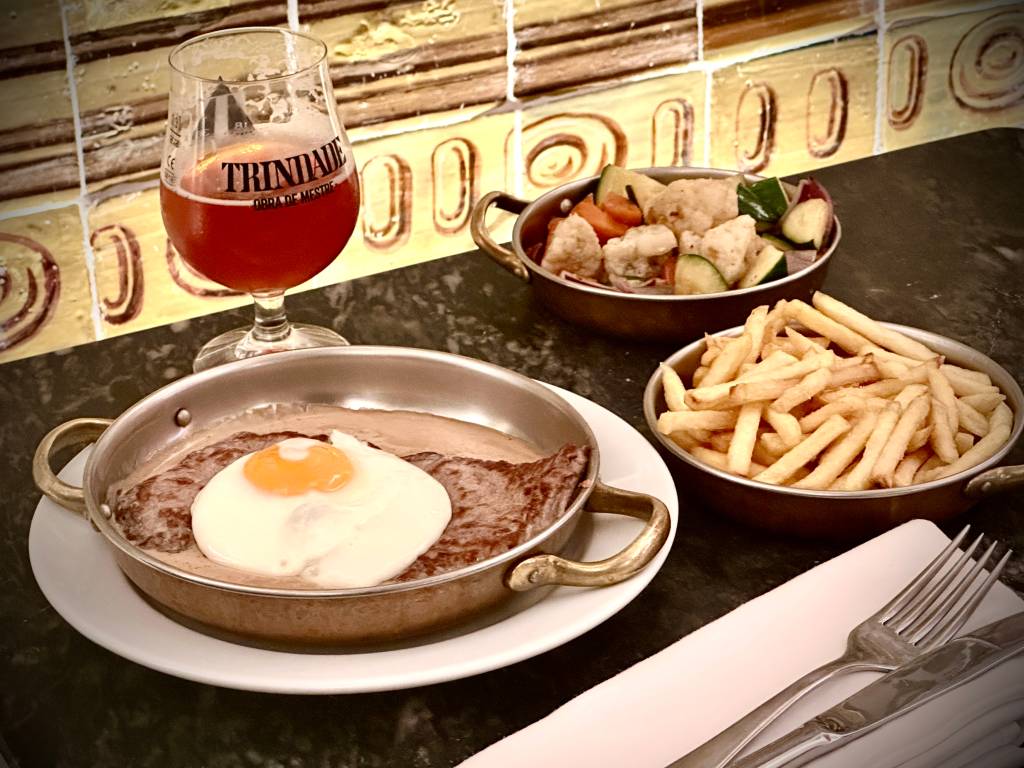 Mesa posta com um prato de bofe e ovo frito, batatas fritas, legumes e um copo de cerveja