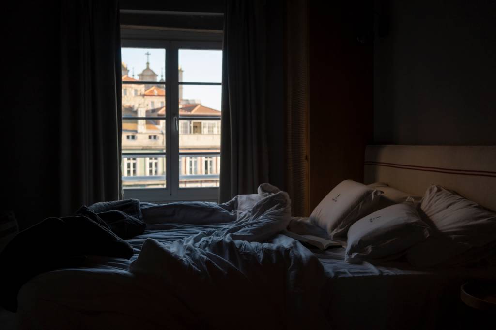 Num primeiro plano, uma cama desarrumada, com uma janela e, do lado de fora, a cidade do Porto