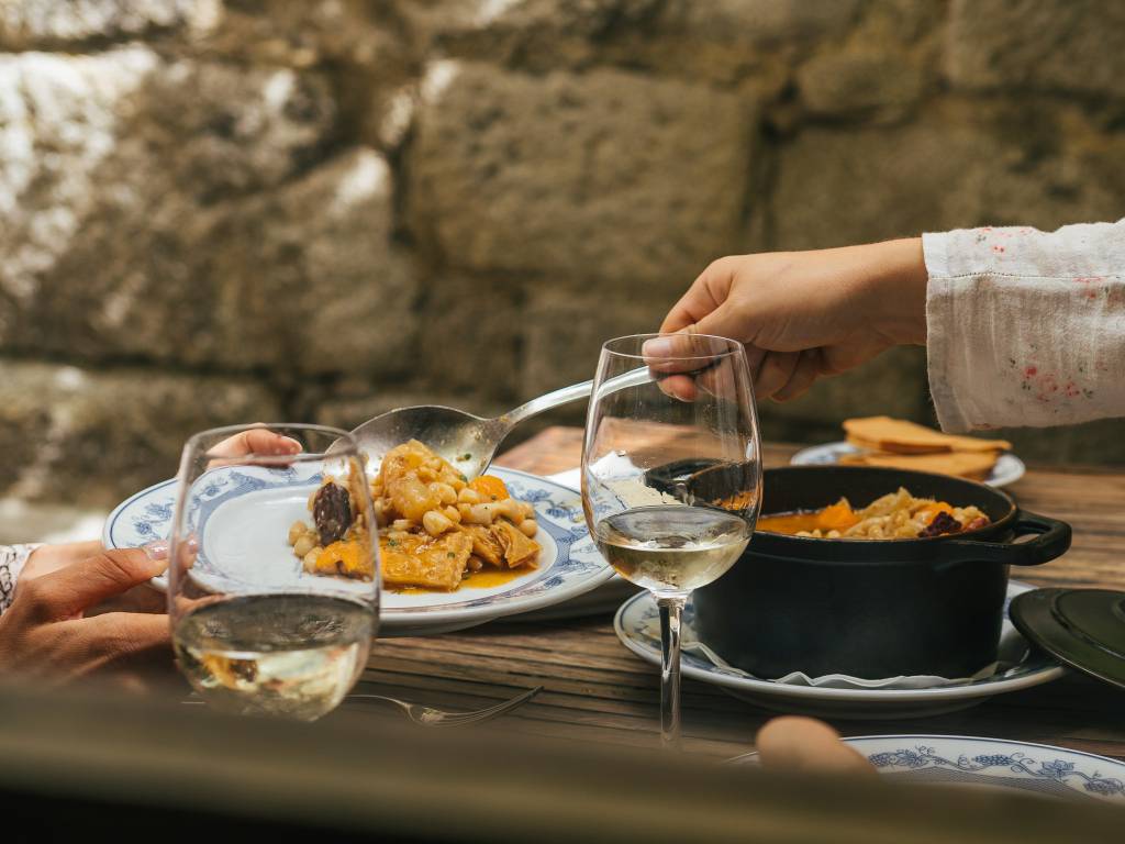 Casal sentado numa mesa ao ar livre para almoçar, servindo-se de um dos pratos, com dois copos de vinho branco ao lado