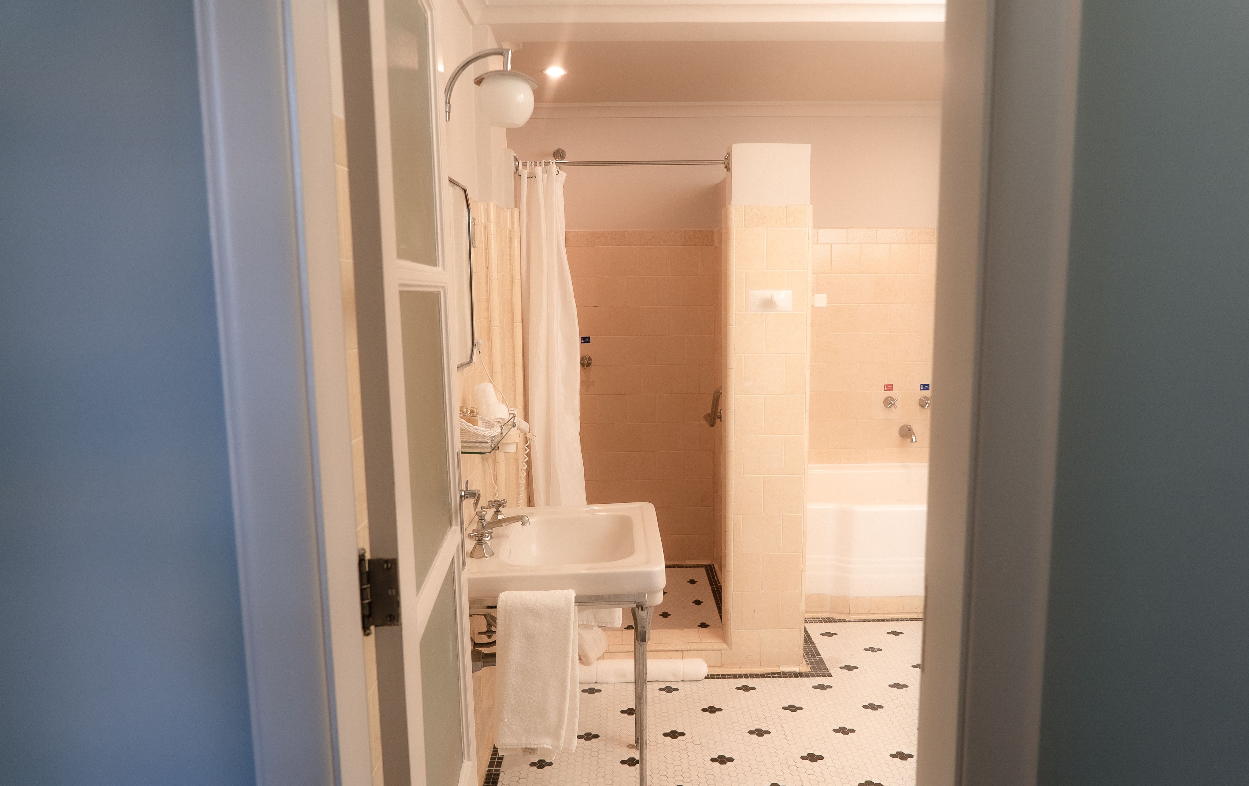 A fotografia colorida mostra um banheiro antigo, com partilhas pretas e brancas no piso e azulejos rosados na parede. Vê-se uma pia, um chuveiro e uma banheira