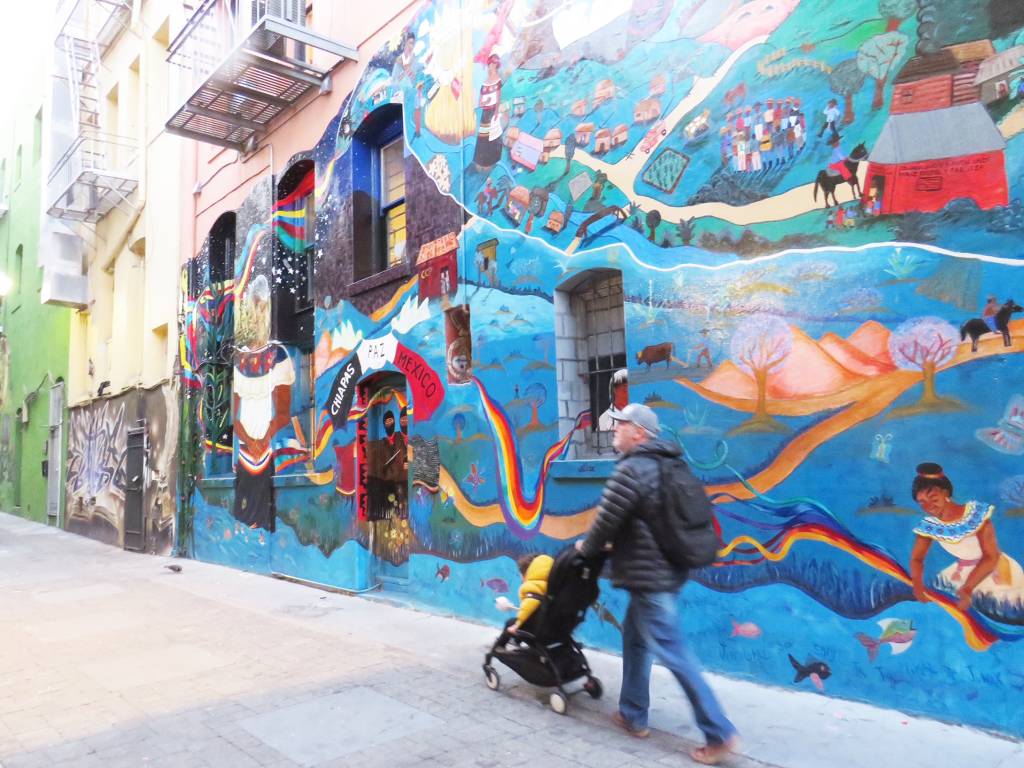 Homem passando com um carrinho de bebê em frente a um mural de grafites