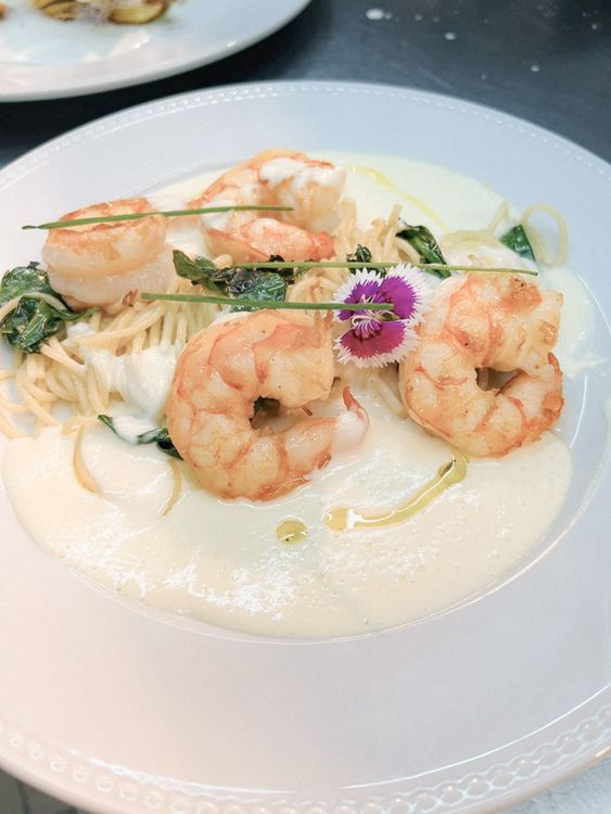 A foto mostra um prato com camarões, macarrão, molho branco e flores comestíveis.