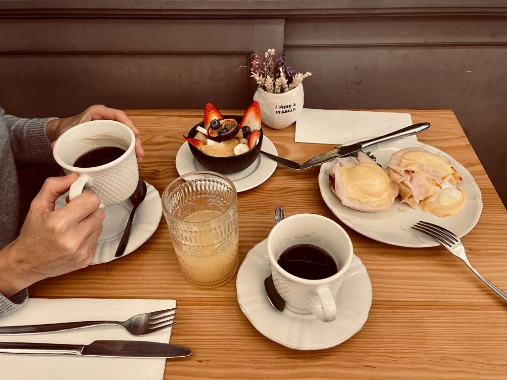 Mesa de café da manhã onde se vê mãos a sgurar uma xícara de café, suco, frutas e ovos