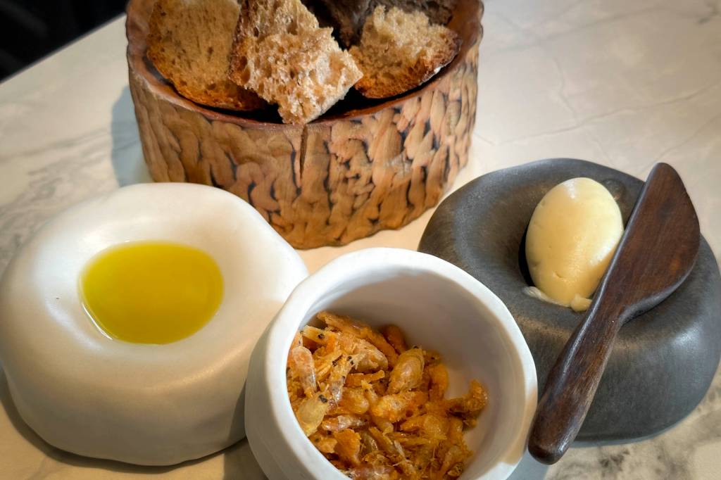 Couvert do restayrante com grossas fatias de pão numa cesta, azeite serviço num bowl, manteiga e um potinho com pequenos camarões fritos