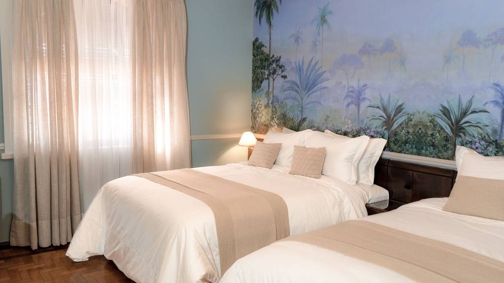 A fotografia colorida mostra um quarto com pinturas florais na parede e duas camas de casal com lençóis brancos.