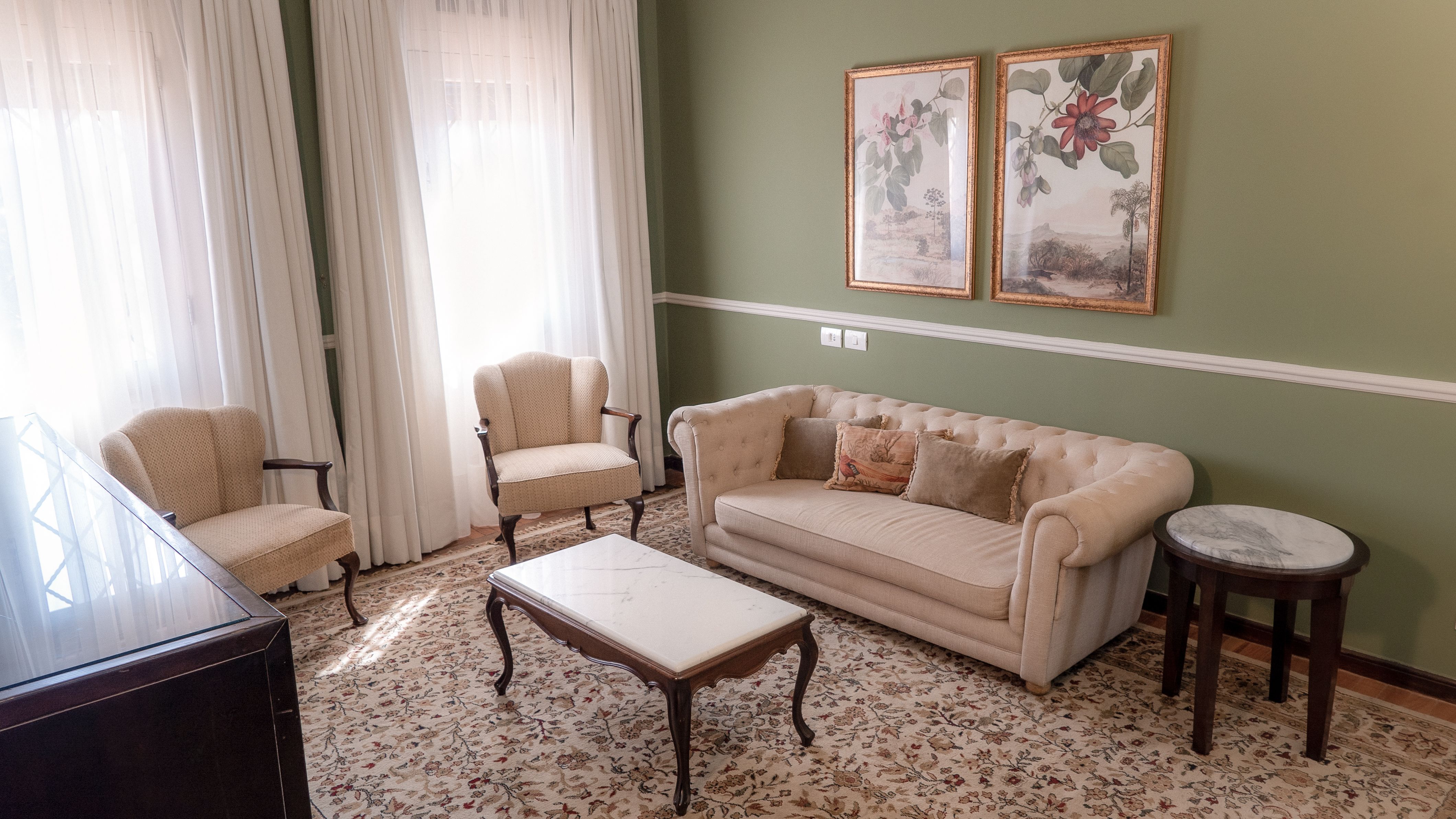 A fotografia colorida mostra uma sala de estar com móveis antigos em creme, parede verde e pinturas florais