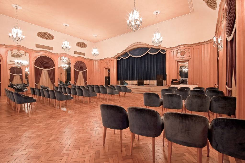A fotografia colorida mostra um salão com paredes de madeira e um palco com cortina escura. As cadeira aveludadas estão distribuídas como uma plateia