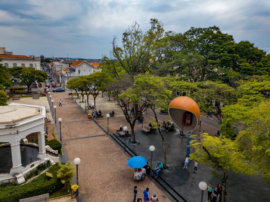 A imagem mostra uma praça com um telefone público gigante e um coreto branco