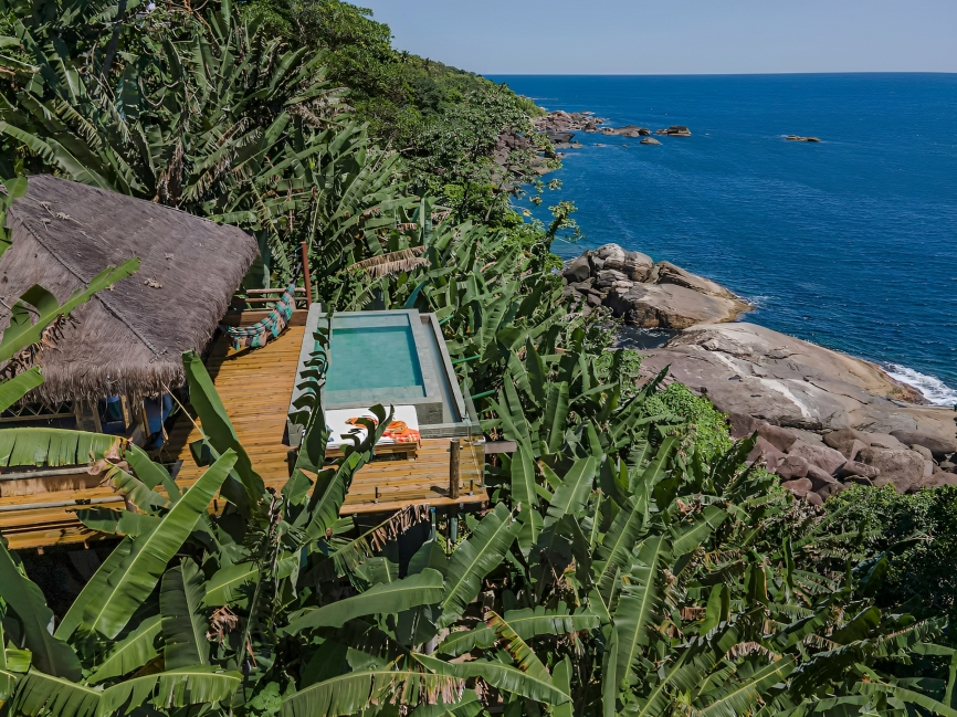 A imagem mostra uma cabana com piscina no meio de uma plantação de bananas, em frente ao mar