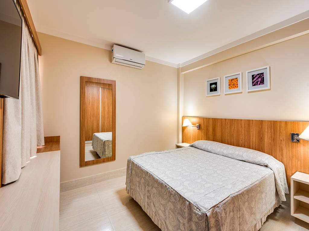 Quarto de hotel com paredes bege, cama de casal com colcha estampada, cabeceira de madeira clara, duas mesas de cabeceira com luminárias, espelho de corpo inteiro, televisão na parede e ar condicionado