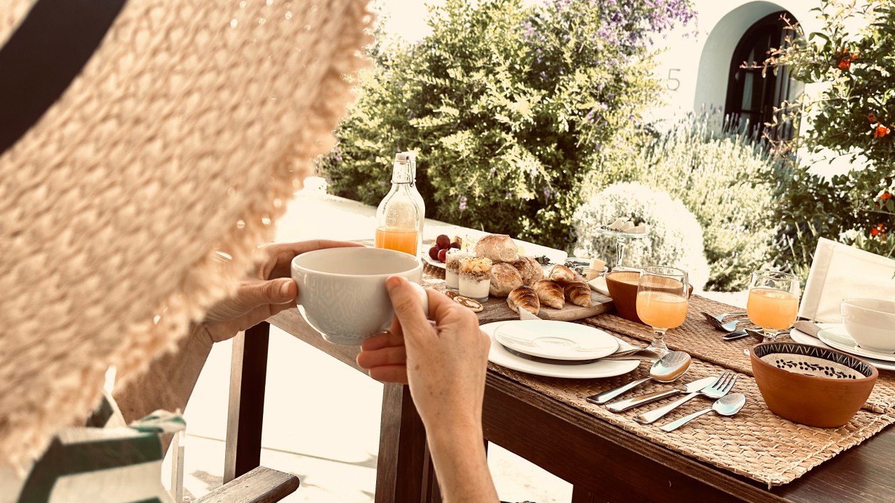 Mulher de chapéu, de costas, sentada à mesa com o café da manhã à frente, segurando uma xícara de café