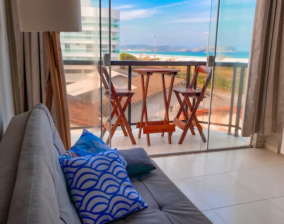Fotografia mostra uma sala com janela de vidro, que dá direto para uma sacada com uma mesa e duas cadeiras com vista para a praia de Cabo Frio