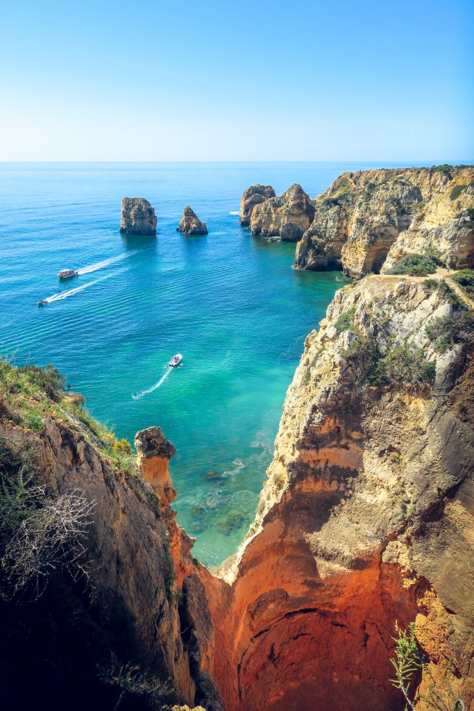 Vista do alto de falésias para um mar em diferentes tons de azul, com barquinhos passando ao fundo