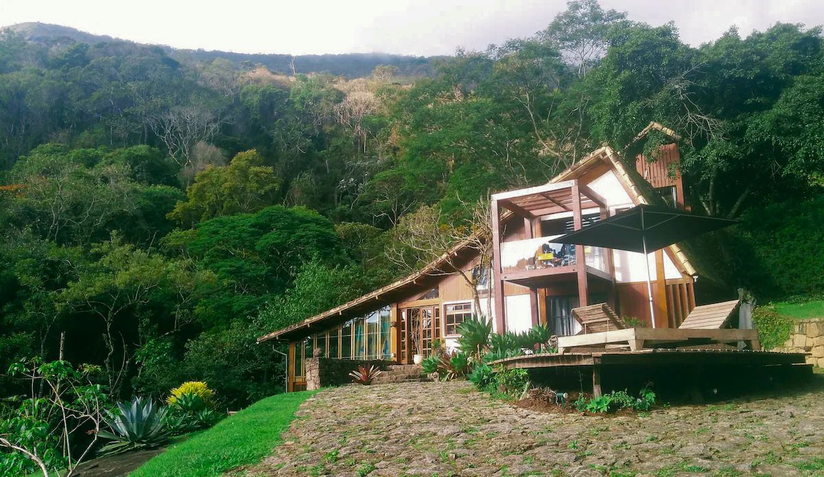 Casa rústica de madeira e pedra com telhado inclinado, varanda e deck com espreguiçadeiras e guarda-sol preto, cercada por mata verde exuberante e céu nublado