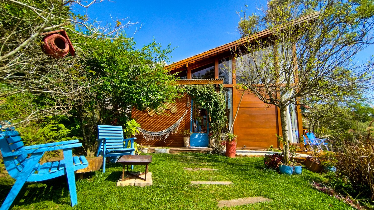Casa de madeira com telhado inclinado e grandes janelas, cercada por gramado verde e árvores. Há duas cadeiras azuis e uma mesa de centro no jardim, com uma rede branca pendurada na varanda. O céu é azul claro