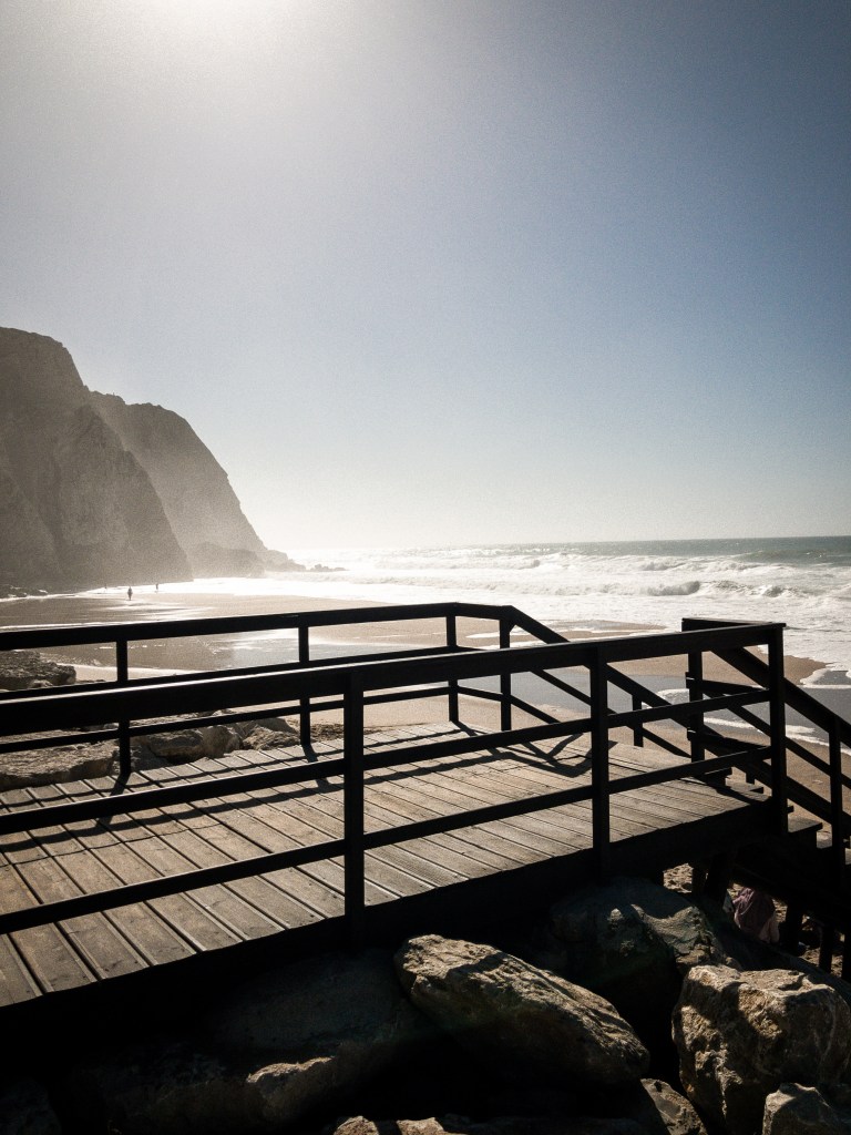 escadaria de madeira que desce para uma praia ao nascer do sol, com falésias ao fundo