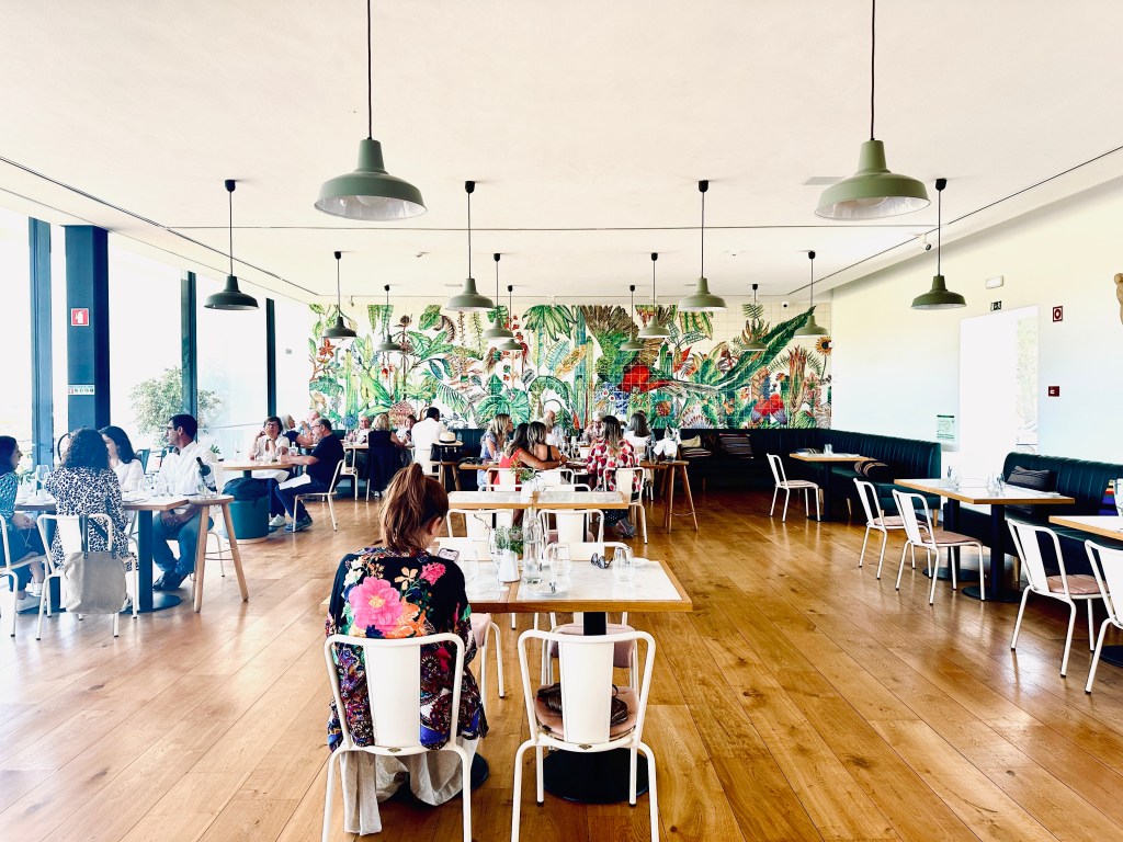 Sala de um restaurante com piso de madeira, pequenas mesas com cadeiras brancas, luminárias pendendes e um grande painel de azulejos coloridos com desenhos de plantas tropicais ao fundo