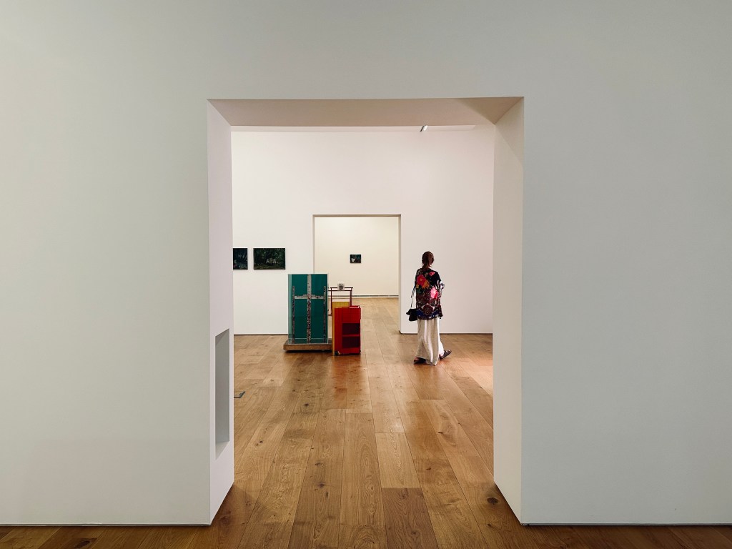 Salas de uma galeria de arte com paredes brancas e alguns quadros coloridos nas paredes, com uma mulher a observá-los