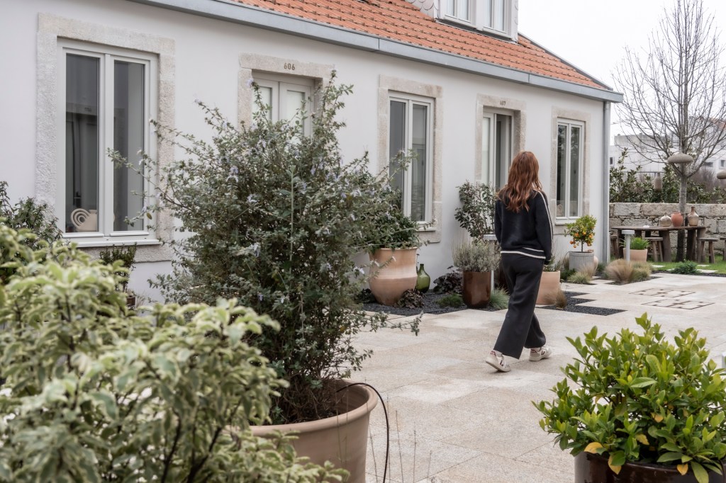 Uma mulher vestida de preto caminha em um pátio em frente a casinhas de fachada branca e plantas