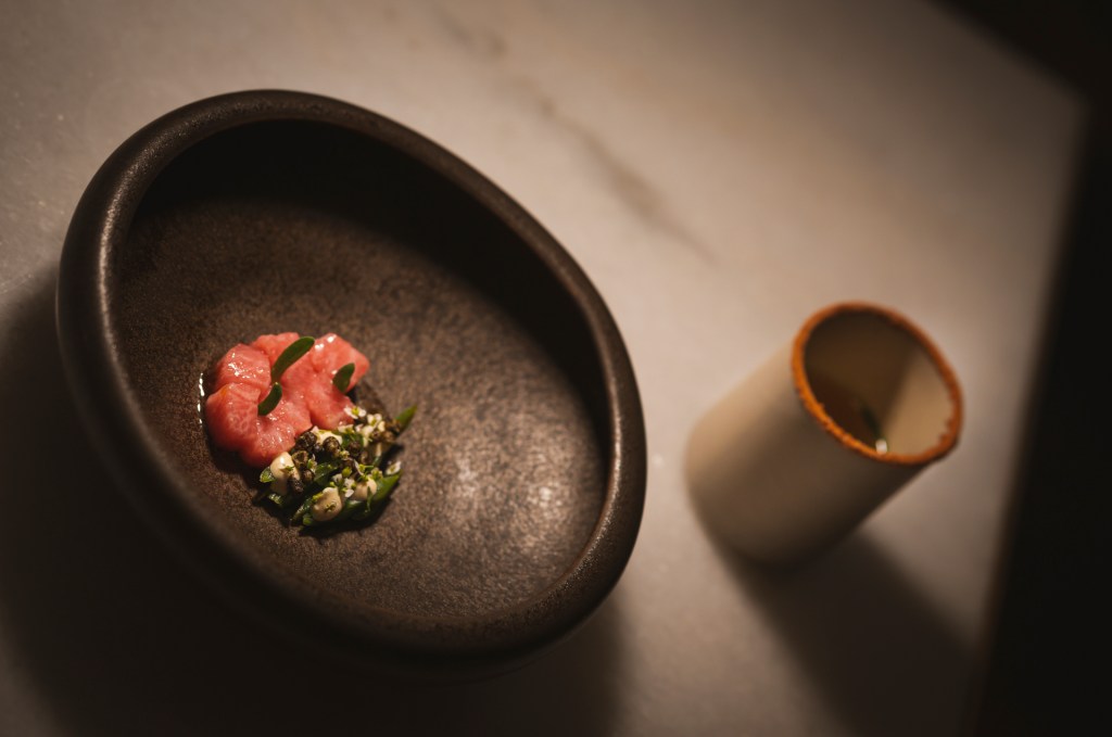 Fotografia colorida. Foto do prato do restaurante. Sobre uma mesa de tampo de mármore branco, um prato de cerâmica cinza com cubinhos de atum e uma guarnição verde