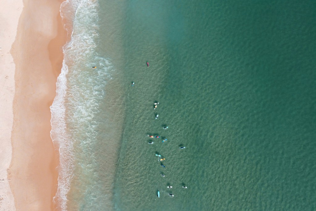 Imagem aérea colorida mostra uma praia com o mar cheio de surfistas a esperar por uma onda