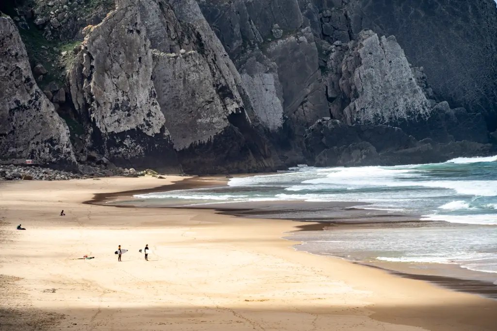 Foto colorida mostra uma praia aos pés de uma imensa escarpa de pedra, com duas pessoas que parecem pontinhos na areia tamanha a imponência das rochas