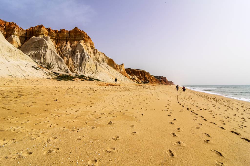 Portugal, Algarve, cliff coast, beach Praia da Falesia Foto colorida mostra o areal de uma praia, onde caminha um casal, com o mar de um lado e falésias coloridas do outro