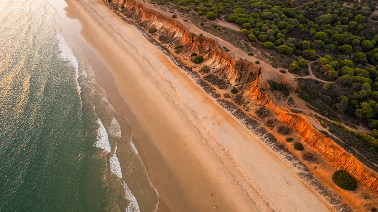Vista aérea de uma foto colorida que mostra uma praia com falésias de cor ocre banhada por um mar de águas azuis
