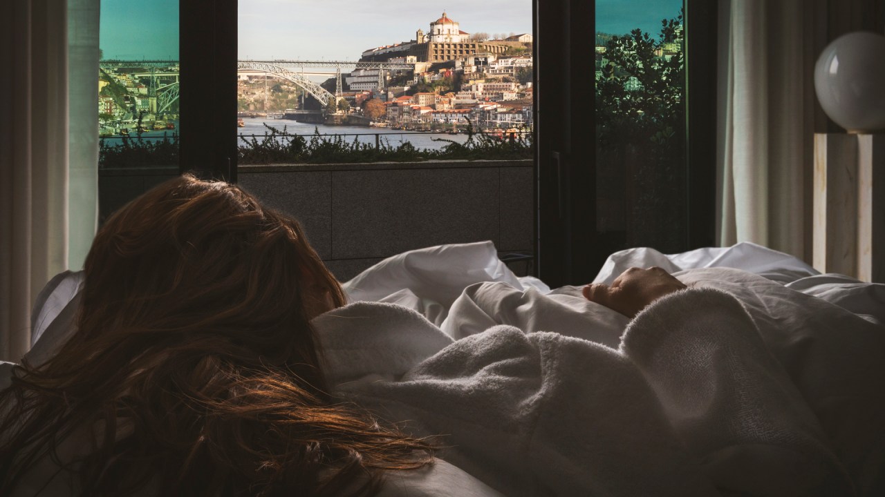Fotografia colorida mostra uma mulher de costas deitada na cama virada para a janela e, do lado de fora, vista do rio Douro, da ponte D. Luis I e do casario colorido de Gaia