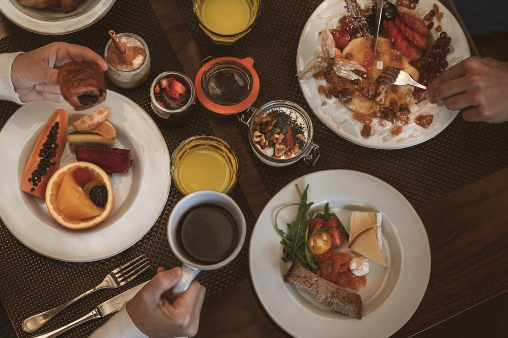 Fotografia colorida mostra em detalhe uma mesa de café da manhã com as mãos de duas pessoas se servindo. Vê-se xicaras de café, suco de laramja, panquecas e frutas