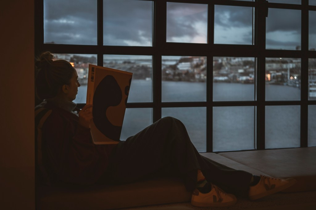 Fotografia colorida mostra uma mulher em primeiro plano sentada ao lado de uma janela lendo uma revista, com a vista do rio e da cidade do Porto do lado de fora