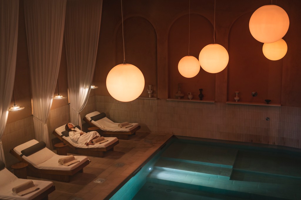 Fotografia colorida mostra a piscina interna de um spa, cercada de espreguiçadeiras, onde uma mulher de roupão relaxa
