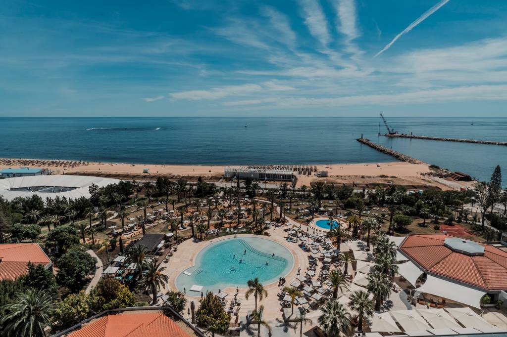 Vista do alto mostra o hotel com uma piscina arredondada cercada de coqueiros, com o mar ao fundo