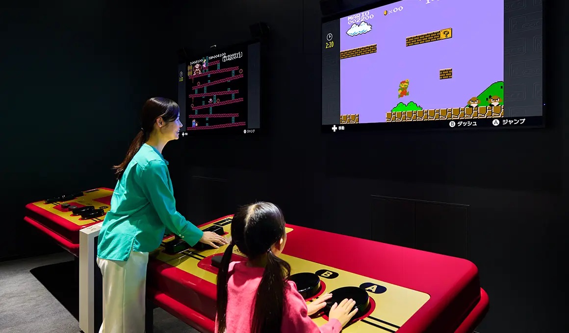 Uma mulher e uma criança se divertem jogando em um controle gigante no Nintendo Museum, Kyoto, Japão