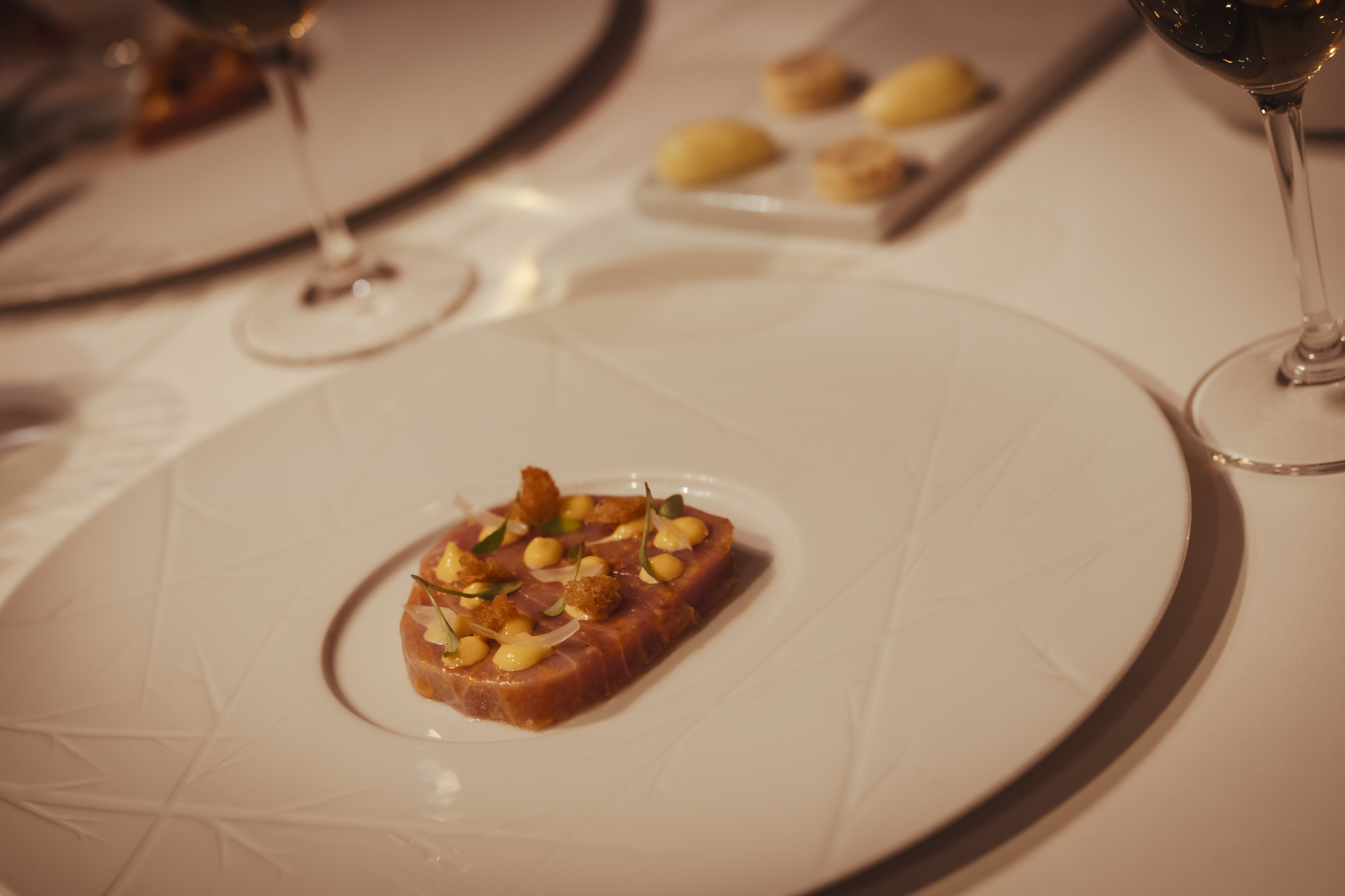 A imagem mostra detalhe de uma mesa no restaurante do hotel, com um prato de carne decorado com bolinhas de molho e flores no topo