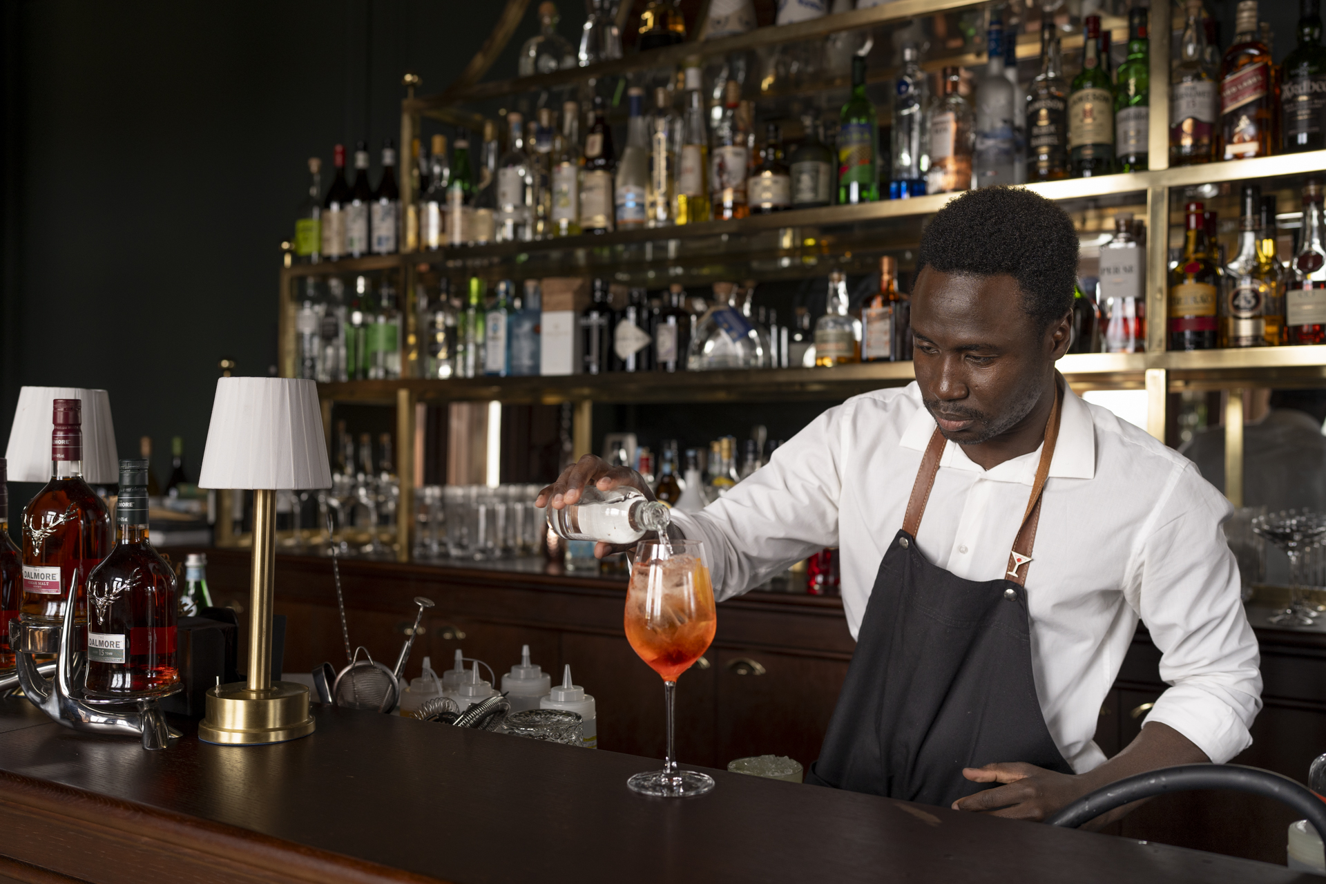 À frente de uma parede repleta de garrafas de bebidas, um barman finaliza um drink de cor laranja no copo