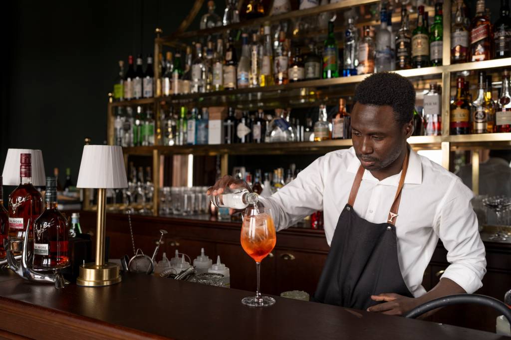 À frente de uma parede repleta de garrafas de bebidas, um barman finaliza um drink de cor laranja no copo