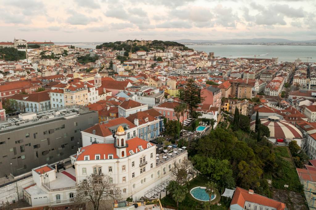 Imagem aérea mostra o casario colorido de Lisboa com destaque para um edifício branco com uma torre e uma piscina redonda, onde fica o hotel Torel Palace Lisboa