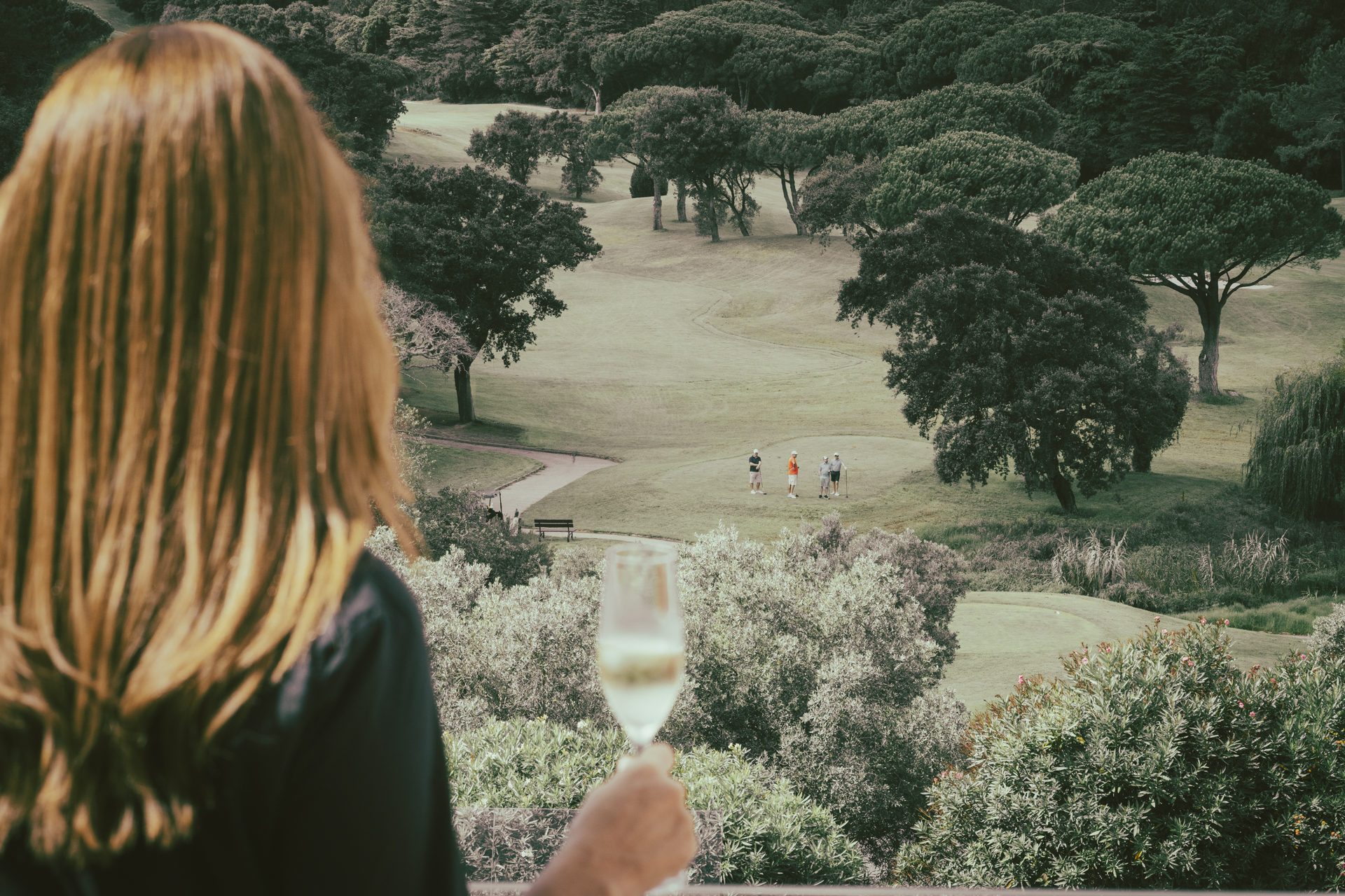 Mulher observa um dos campos de golfe do hotel da varanda de sua suíte, com uma taça de espumante na mão