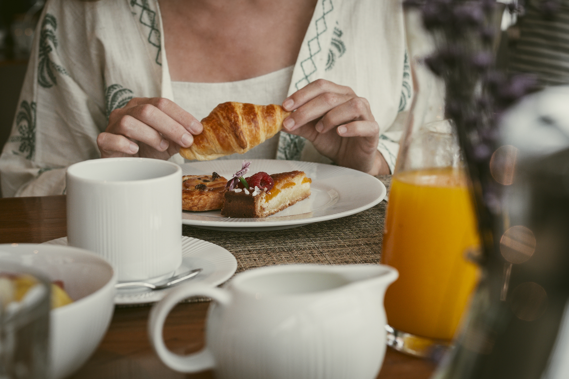 Detalhe de uma mesa de café da manhã do hotel: hóspede a segurar um croissant, com um copo de suco de laranja à frente