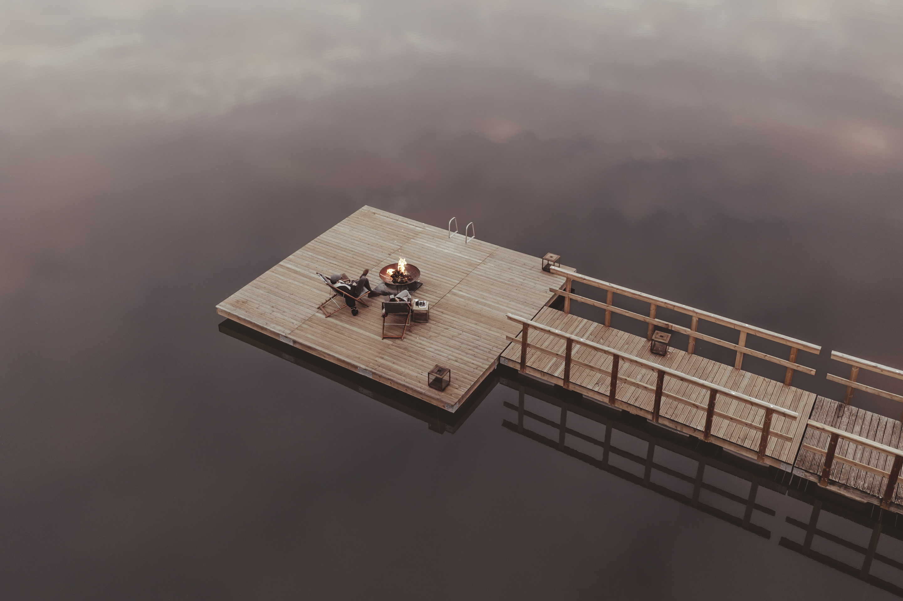 Imagem aérea mostra um deck de madeira sobre o lago do hotel, com um casal sentado em duas cadeiras em volta de uma fogueira